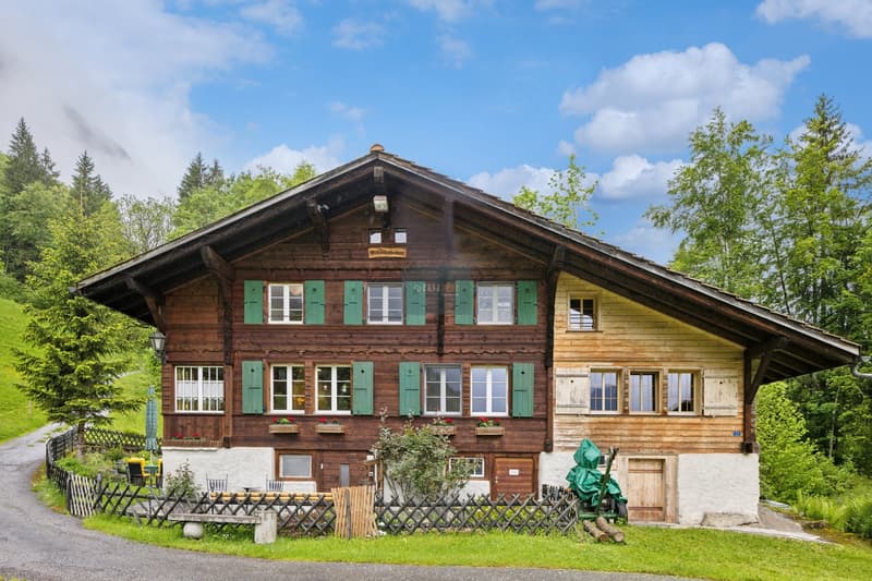 Two-story wooden chalet-style house with green shutters, surrounded by trees and greenery, with a balcony or terrace visible.