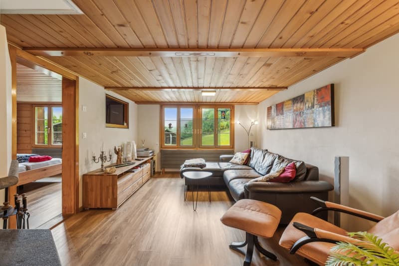 Spacious living room with wooden ceiling and walls, large windows overlooking a scenic outdoor view, leather sofa, wooden entertainment unit, and artwork on the walls.