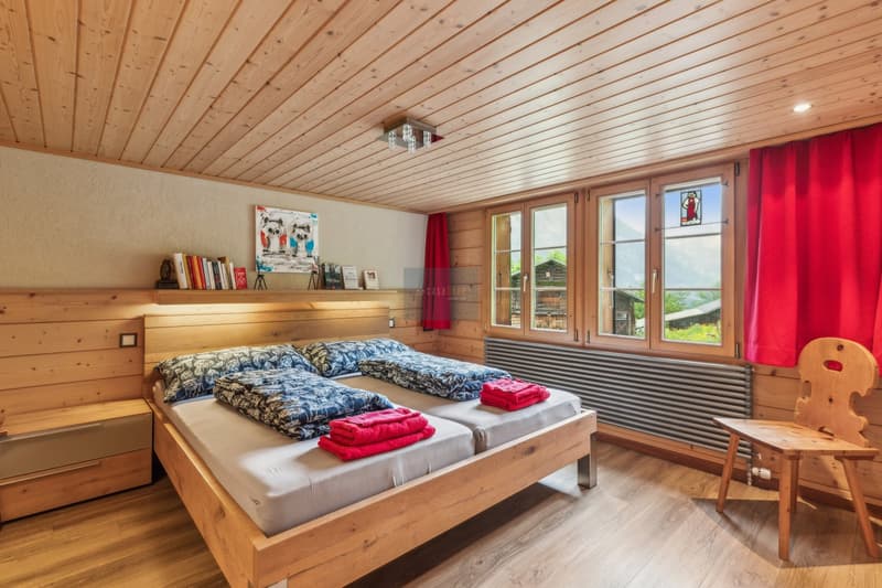 Spacious bedroom with wooden walls and ceiling, large windows providing natural light, a king-size bed with patterned bedding, red accent pillows, and a wooden chair. There is also a shelf with books and decorative items, and a stained glass window panel