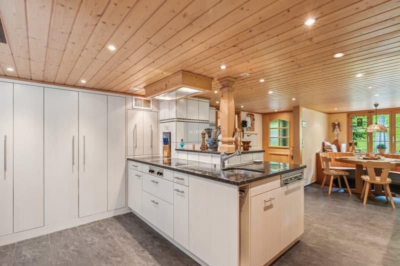 Spacious kitchen with wood-paneled ceiling, white cabinets, granite countertops, and a kitchen island with a sink and dishwasher. The kitchen opens up to a dining area with a wooden table and chairs.
