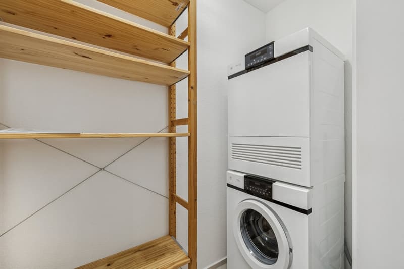 The image shows a utility room with a washing machine and dryer unit, as well as wooden shelving units for storage.