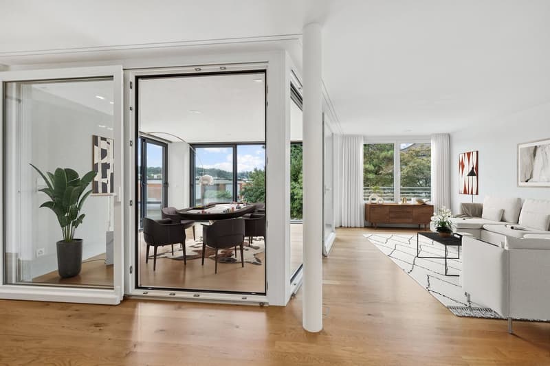 Spacious open-plan living area with large windows, hardwood floors, modern furniture including a dining table and chairs, and a living room with a sofa and coffee table. The room has access to a balcony or terrace with a view of the surrounding greenery.