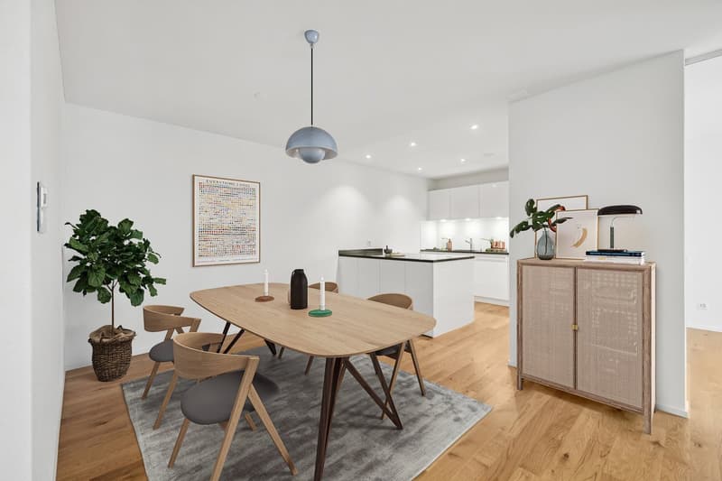 The image shows an open-plan living area with a dining table, chairs, and a kitchen. The dining table has a wooden top and dark legs. The chairs around the table are also wooden with a natural finish. The kitchen has white cabinets and a black countertop.
