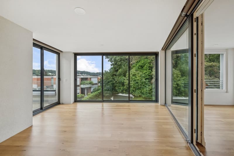 Spacious living room with large windows offering a scenic view of the surrounding greenery, hardwood flooring, and sliding glass doors leading to a balcony or terrace.