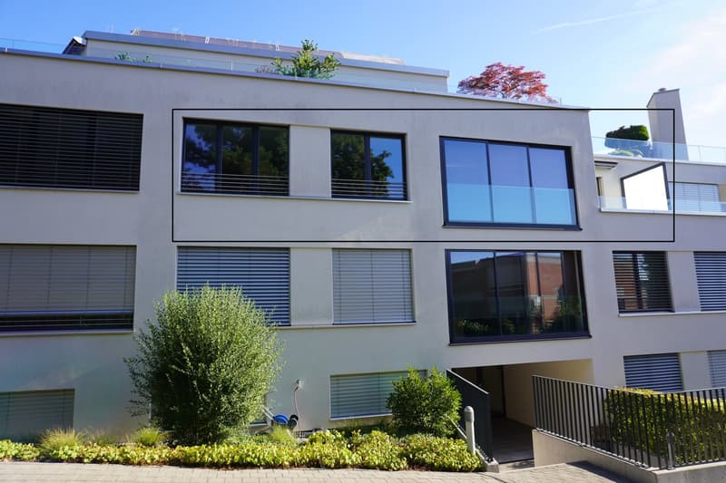Modern multi-story apartment building with large windows, balconies, and landscaped gardens in the front.