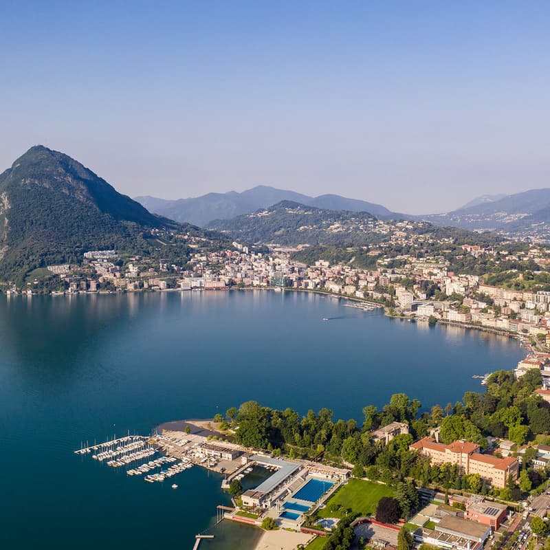 This image shows an aerial view of a scenic lakeside town surrounded by mountains. The town is situated along the shores of a large, blue lake with a marina and boats docked. The town appears to be built on the slopes of the surrounding hills, with a mix