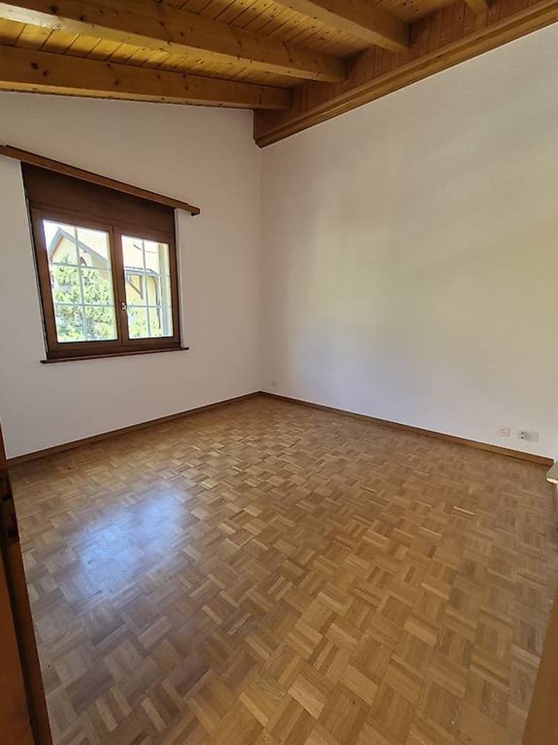 Empty room with wooden parquet flooring, wooden ceiling, white walls, and a large window.