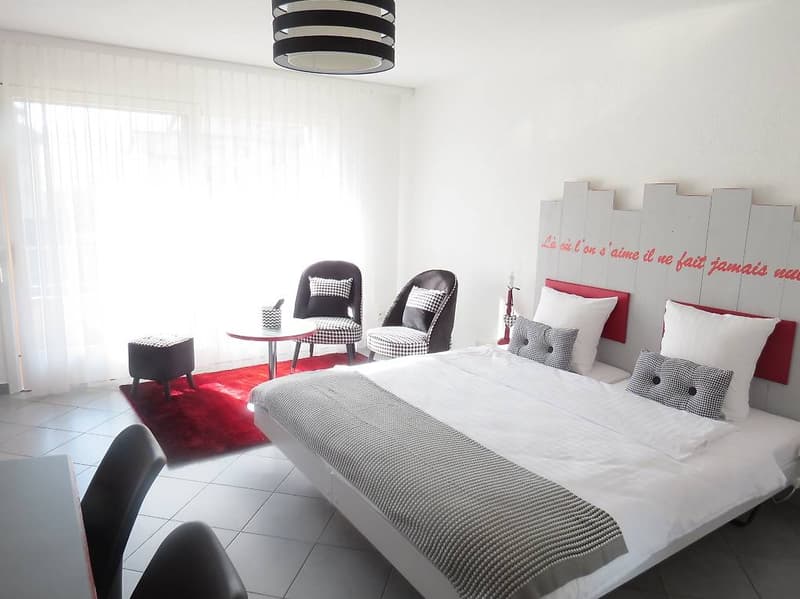Large bed, white bedding, red rug, white walls, white curtains, stools, chairs, table, lamps, wooden headboard with French text