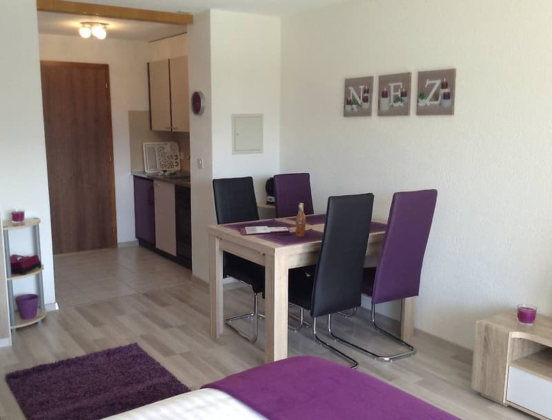 dining area with table and 4 chairs, bed with purple blanket, wooden shelves on the wall, small desk, kitchen, carpet