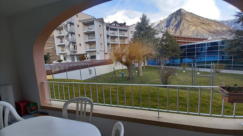 balcony, table, chairs, metal railing, green grass, trees, building, mountains