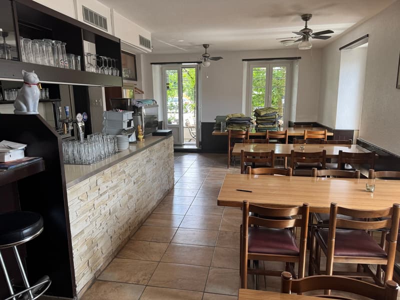 The image shows an open-concept kitchen and dining area. The kitchen features a stone-tiled counter with a built-in dishwasher, shelves displaying glassware, and a coffee maker. The dining area has a wooden table with chairs, and there are additional cush