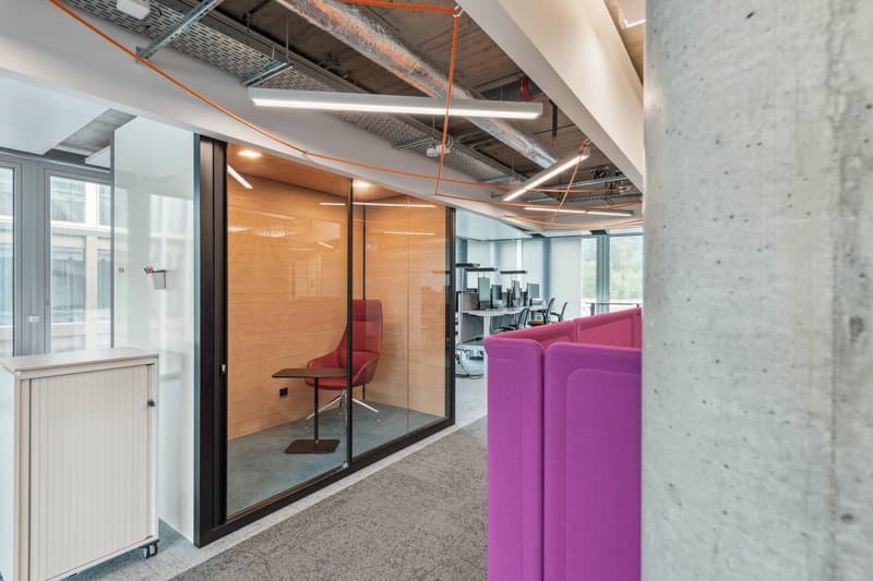 open glass walled room, purple lounge seating, desk with red chair, white office storage unit, office chairs and desks in the background