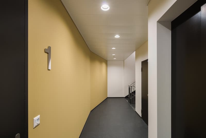 Corridor with yellow wall, black floor, black doors, white ceiling with recessed lighting, power outlet, stairs leading up, hallway numbering.