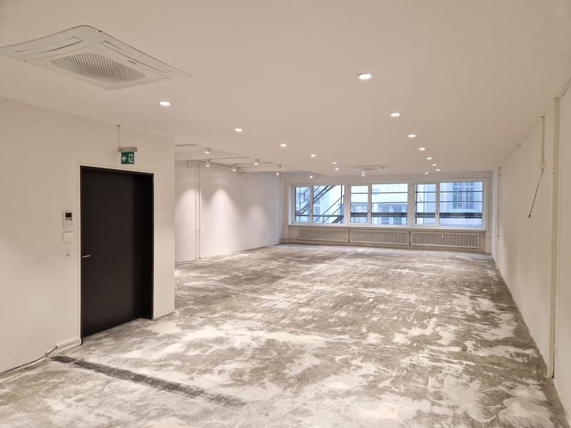 Empty room with white walls, gray floor, large windows, and a door.