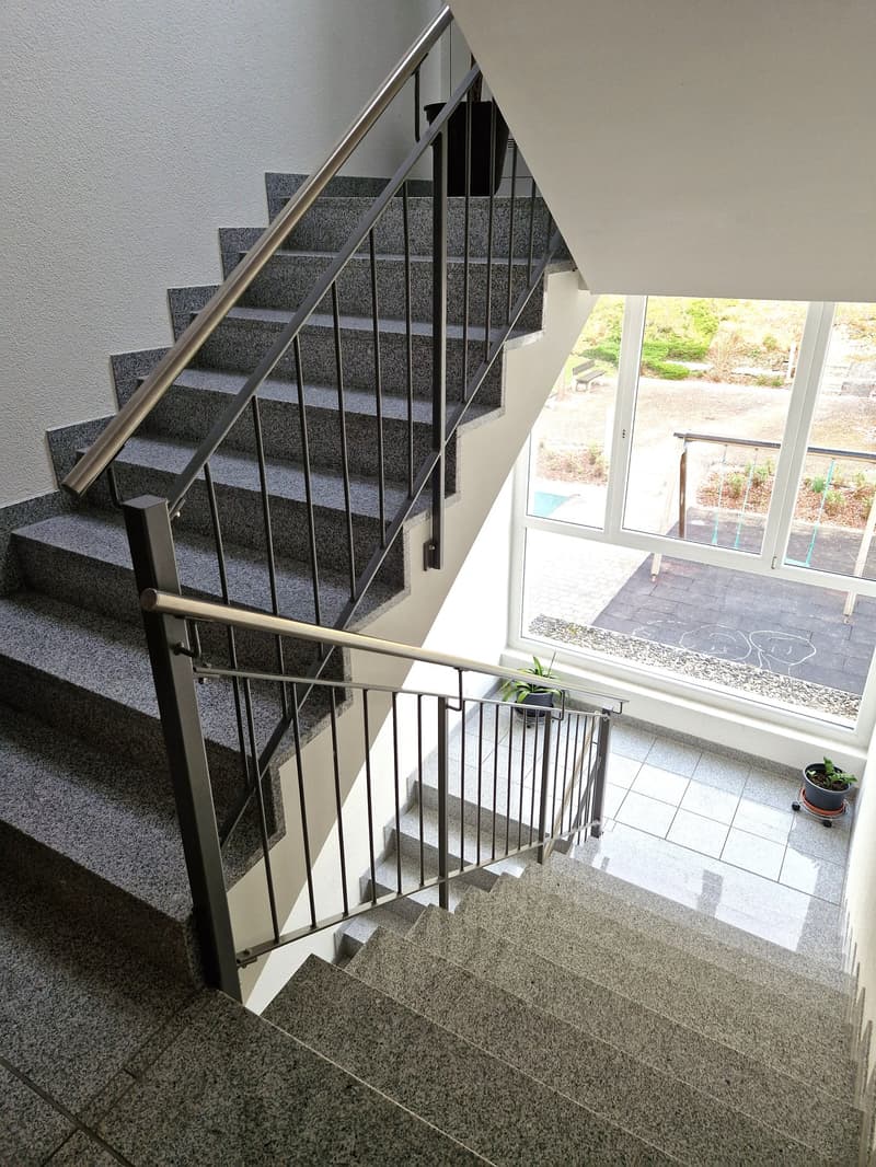 The image shows a staircase with granite steps and a metal railing. The staircase leads to a balcony or terrace visible through the large windows at the top.