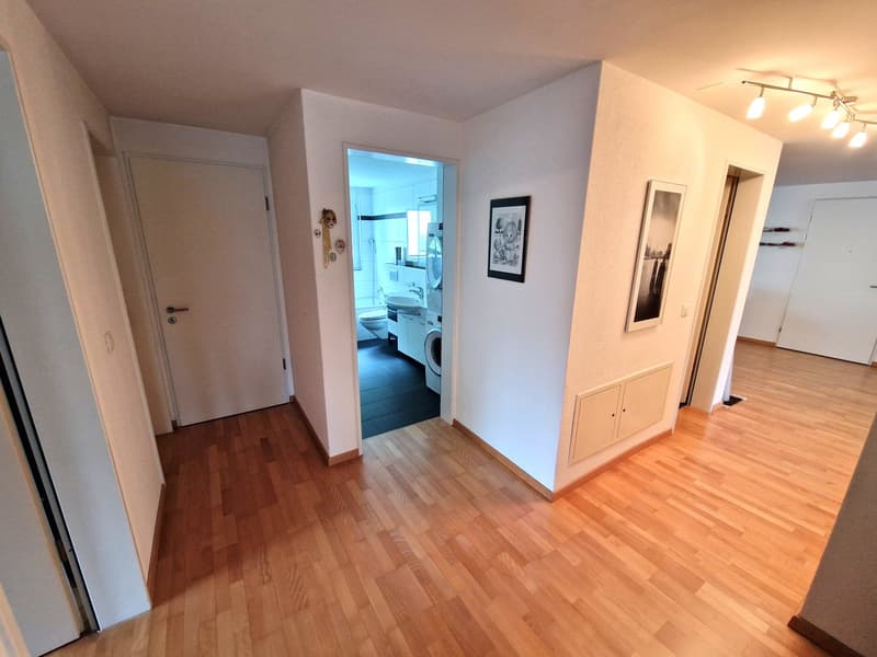 The image shows the interior of an apartment. The main room has hardwood floors and white walls. There is a doorway leading to another room, which appears to be a bathroom based on the visible fixtures. The apartment also has a built-in cabinet or storage