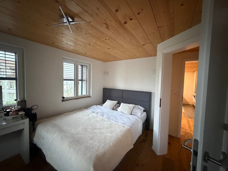 Bedroom with wooden ceiling, a bed, a desk with items, a window, and an open door