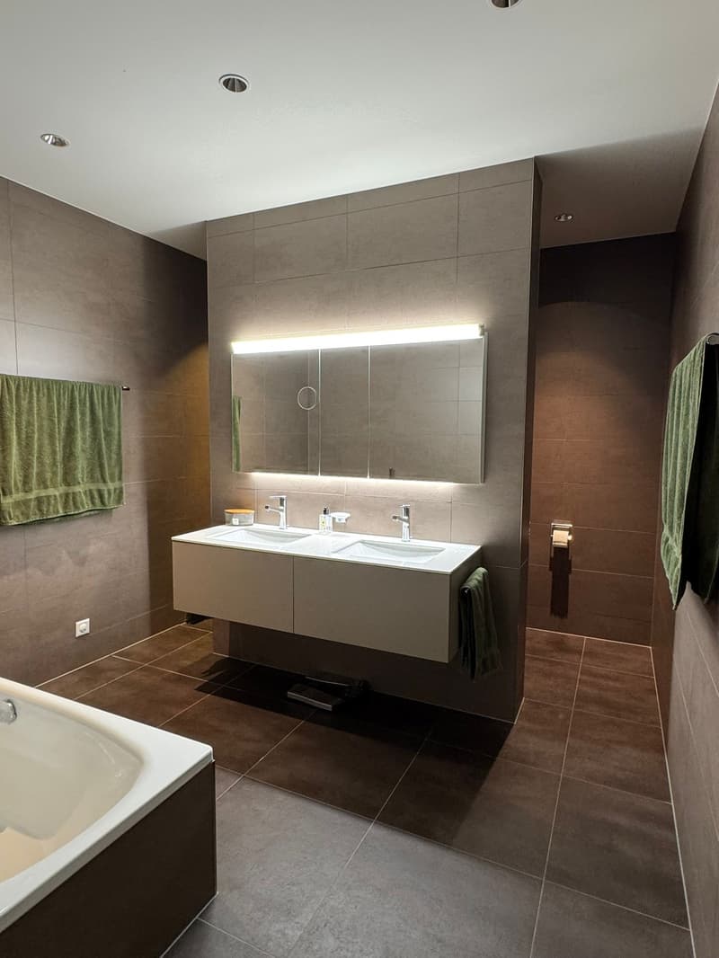 Bathroom: White bathtub, Double sinks, mirror, two green towels, lighting above mirror, brown tiled floor.