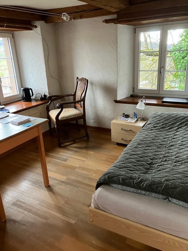 Bedroom with bed, desk, chair, window, wooden floor, nightstand with lamp, radiator, lamp, power outlet, kettle, cup, papers, books, laptop, magazines, bottle, and a toy duck.