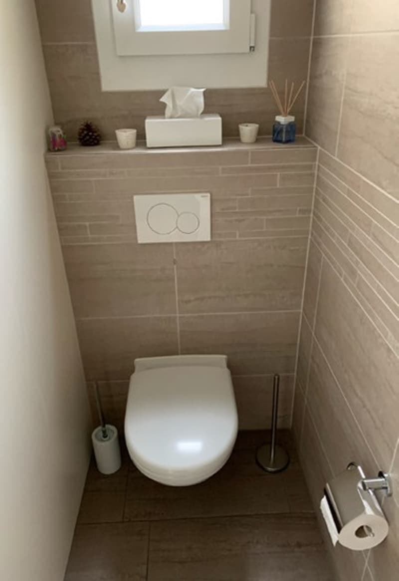 Modern, compact bathroom with beige tiles, white toilet, window with natural light, roll of toilet paper on holder, toilet paper on holder, tissue box, and small items on shelf