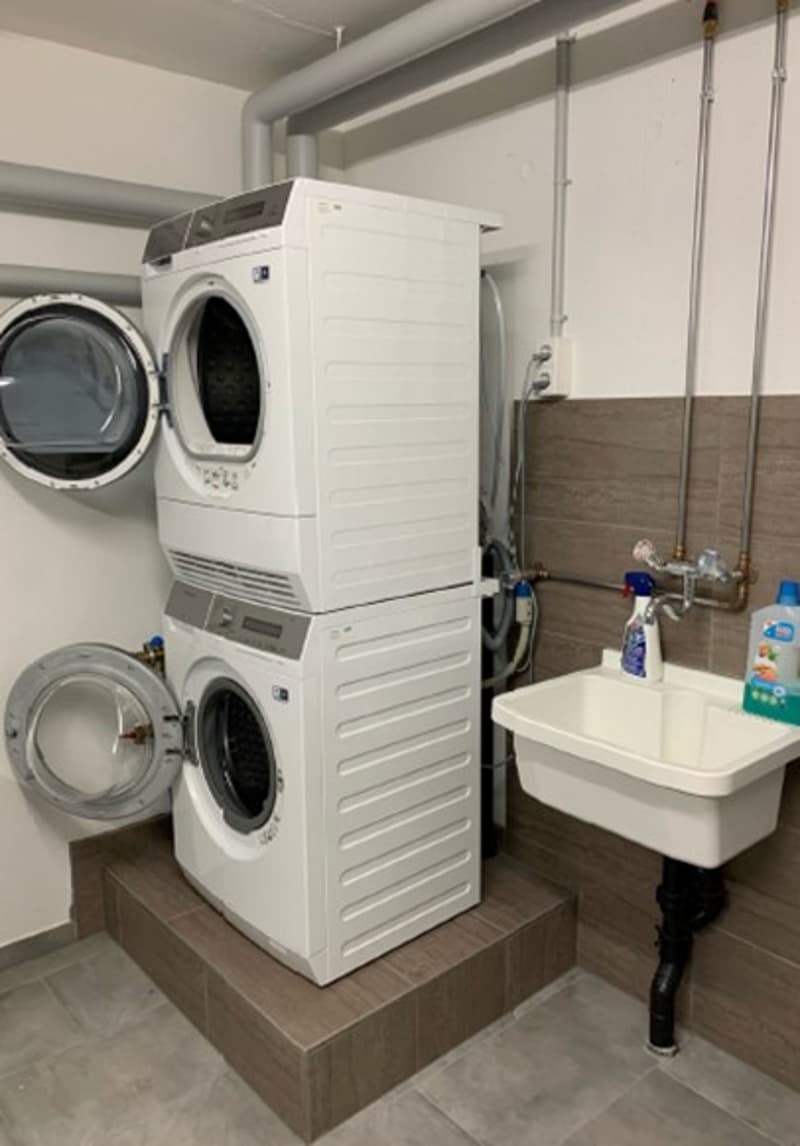 Stacked washer and dryer, sink with faucet, cleaning supplies on the wall, tiled floor, wooden shelf