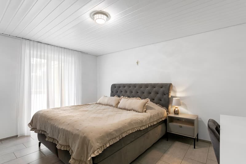 double bed, gray tufted headboard, white walls, large windows with white curtains, nightstand, lamp, chair
