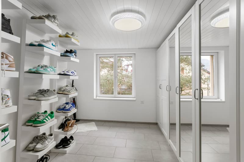 A clean and tidy room with a bright white ceiling and walls. It features shelving units filled with various shoes on the left side, along with two large windows that allow ample natural light to enter. There are also two white doors, one of which seems to