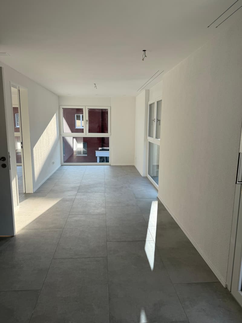 Empty room with white walls, gray tile flooring, large windows, and doors.