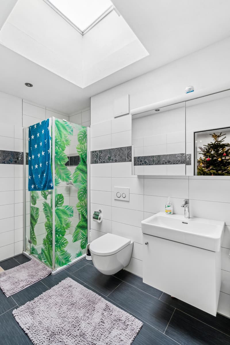modern bathroom with white tiles, glass shower enclosure, white toilet, white sink, white vanity, black floor, white ceiling with skylight, towel, and Christmas tree in mirror
