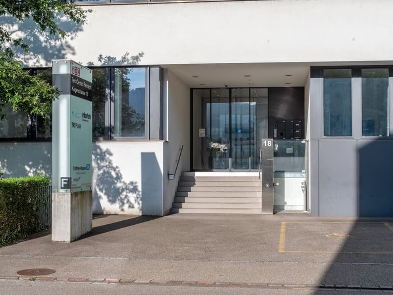 Modern building exterior with glass doors, large windows, concrete walkway, sign, staircase leading to entrance, greenery, and gray color scheme.