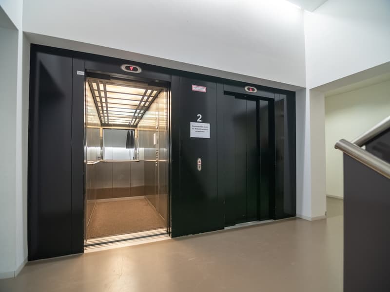 An elevator with glass doors and an illuminated interior. The number '2' is displayed above the door. The elevator's floor is carpeted, and the surrounding walls are painted white.