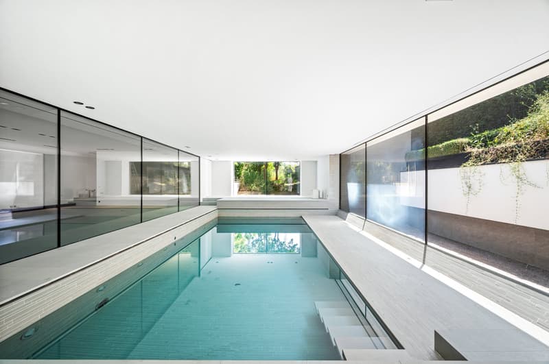 Large indoor swimming pool with glass walls and doors, providing a view of the surrounding greenery. The pool has a minimalist, modern design with a tiled floor and steps leading into the water.