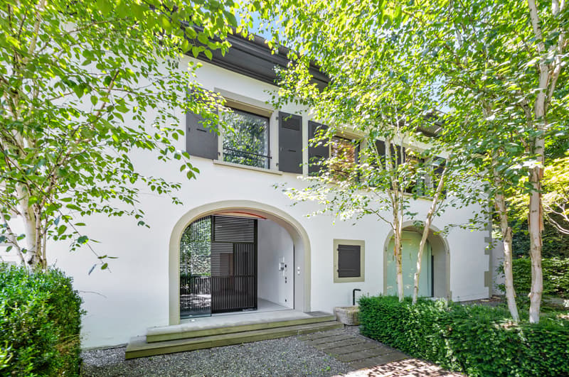 Two-story white house with arched entryway, surrounded by lush greenery and trees, with balconies on the upper level