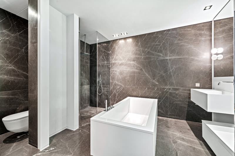 Large bathroom with dark gray marble-like tiles covering the walls, a freestanding white bathtub, a glass shower enclosure, and a vanity with a sink and mirror.