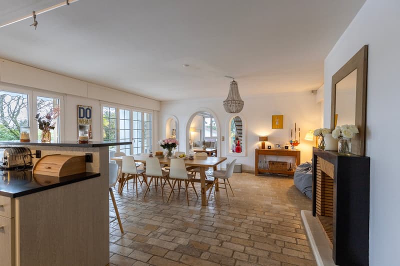 large dining room with brick flooring, a table and chairs, a chandelier, a fireplace, large windows, wooden table, lamp