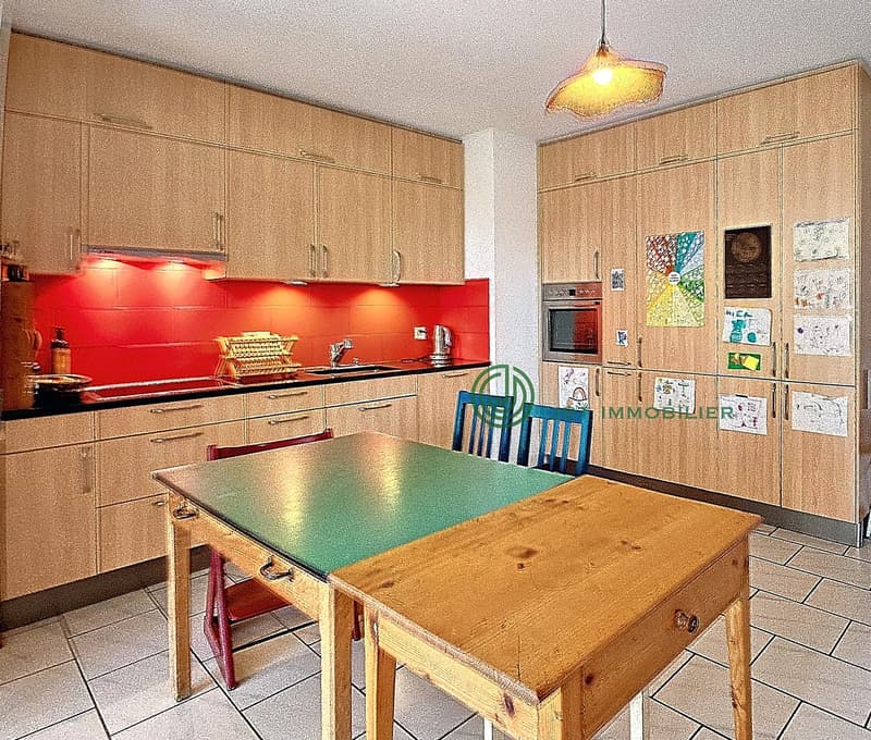 a large room with tiled floor, wooden tables, chairs, cabinets, counter with sink, red accent wall, built-in oven, microwave, cabinet door with pictures