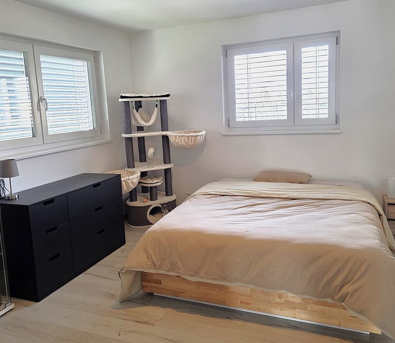 Bedroom with bed, nightstand, lamp, dresser, cat tree, and windows with blinds