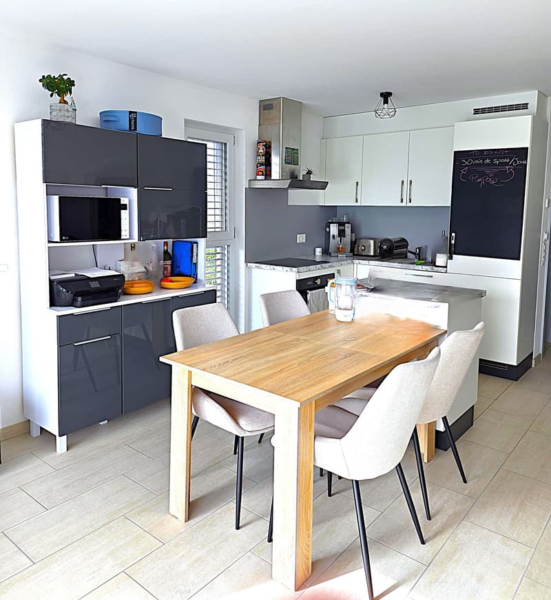 modern kitchen with dining area, wooden table, white chairs, white cabinets, black microwave, printer, blackboard, potted plant, wine bottle, toaster, coffee machine, kettle, sink, gas stove