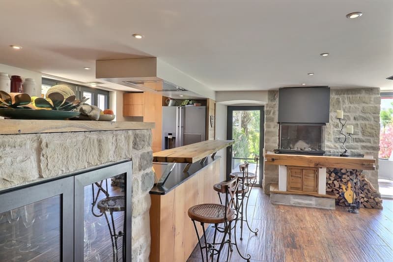 Open kitchen with large island, granite counter tops, modern appliances, fireplace, stools, wooden flooring.
