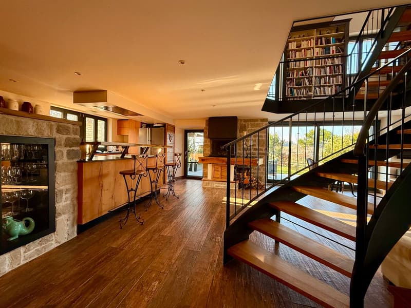 Spacious open floor plan, hardwood floors, large fireplace, spiral staircase with railing, open kitchen, breakfast bar, wooden shelves with books, wine glasses displayed, spiral staircase leading to upper level, balcony, large windows, view of landscape.