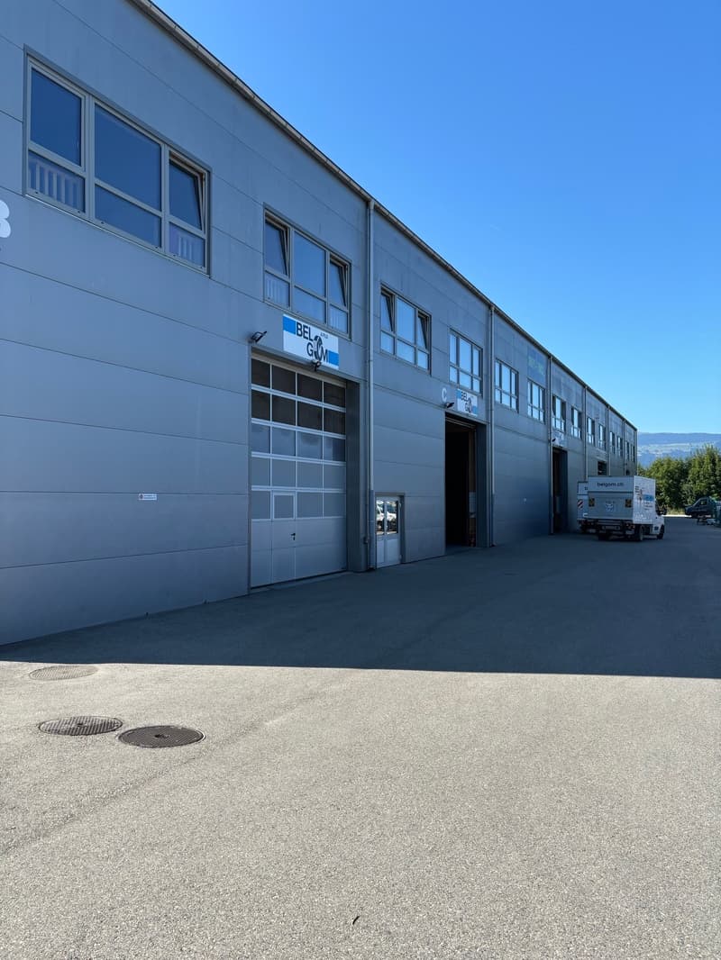 Large industrial or commercial building with multiple garage doors and windows. The building has a gray exterior and is situated on a paved lot.