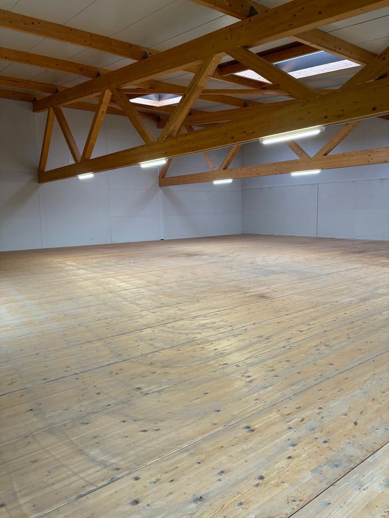 Large open space with wooden beams and trusses in the ceiling, wooden floor, and white walls.