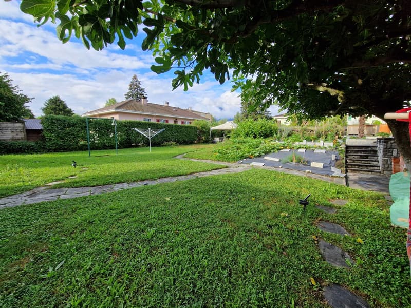 Two-story house with a red tile roof, surrounded by a well-maintained garden with a grassy lawn, trees, and a paved walkway. There is a garage and parking space visible on the property.