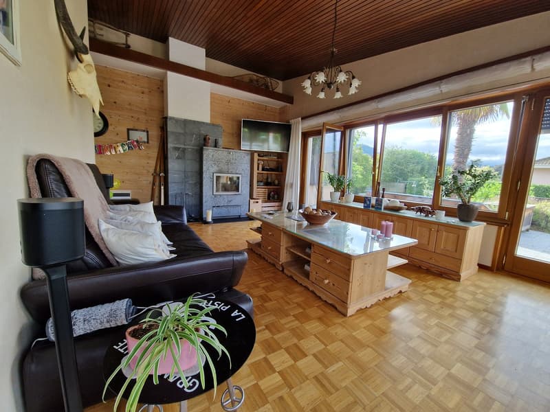 Spacious living room with wooden ceiling, large windows providing a scenic view, leather sofa, wooden cabinets and shelves, and a stone fireplace