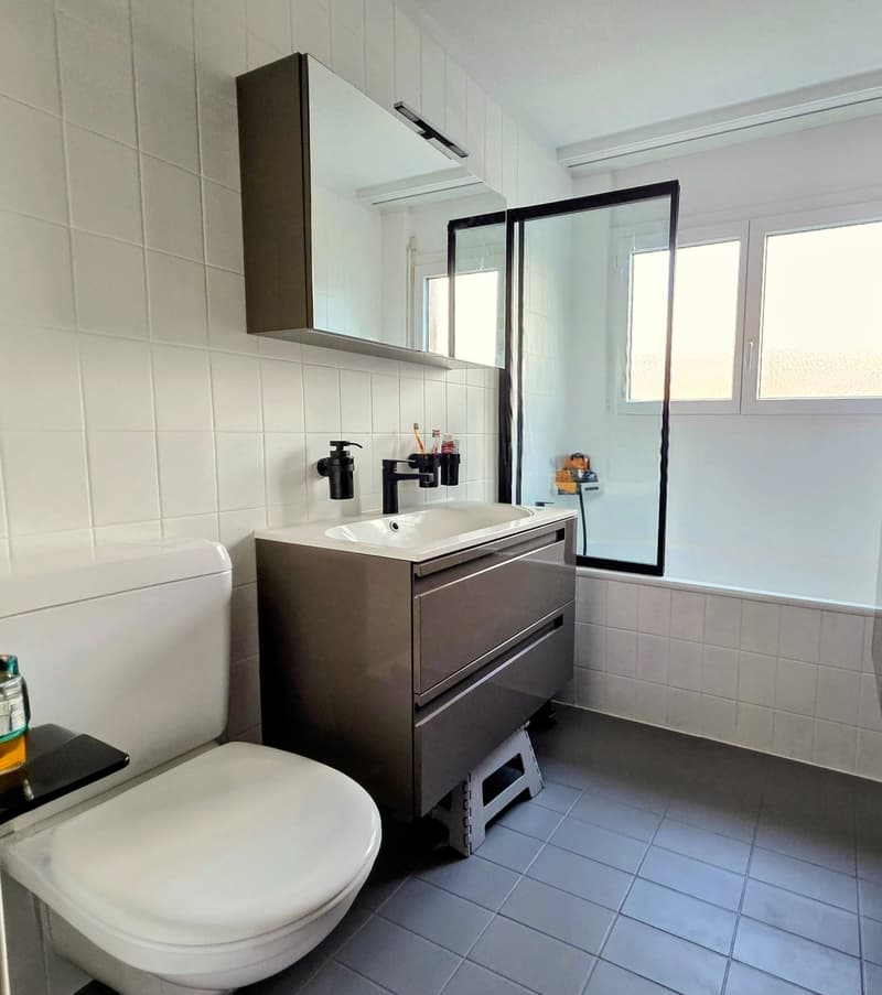 White tiled bathroom, sink with faucet, toilet, large mirror, bathtub with shower, shelves, black bottle, and toothbrush holder