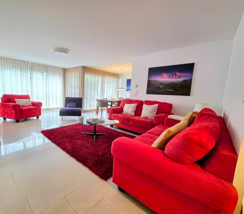 Living room with red couches, red carpet, glass coffee table, white tile floor, white walls, large windows with curtains, picture frame on the wall, and a lamp.
