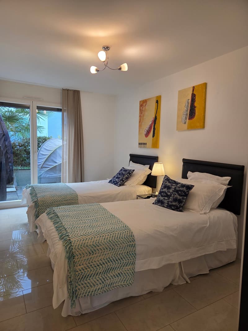 two twin beds, white sheets, blue and white throw pillows, bedside tables with lamps, paintings on the wall, sliding glass door with curtains, tiled floor