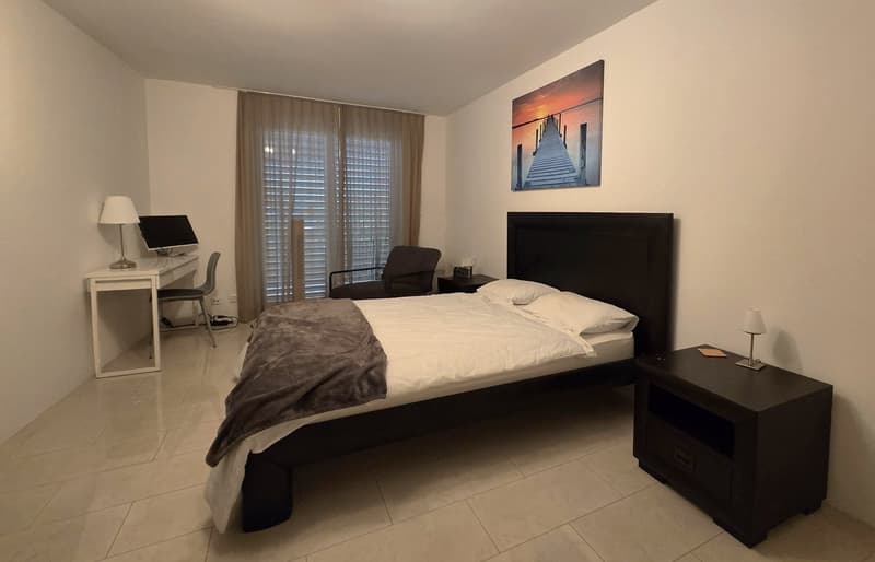 a neatly furnished bedroom featuring a bed, nightstands, a desk with a chair, a painting, and ample window space with blinds