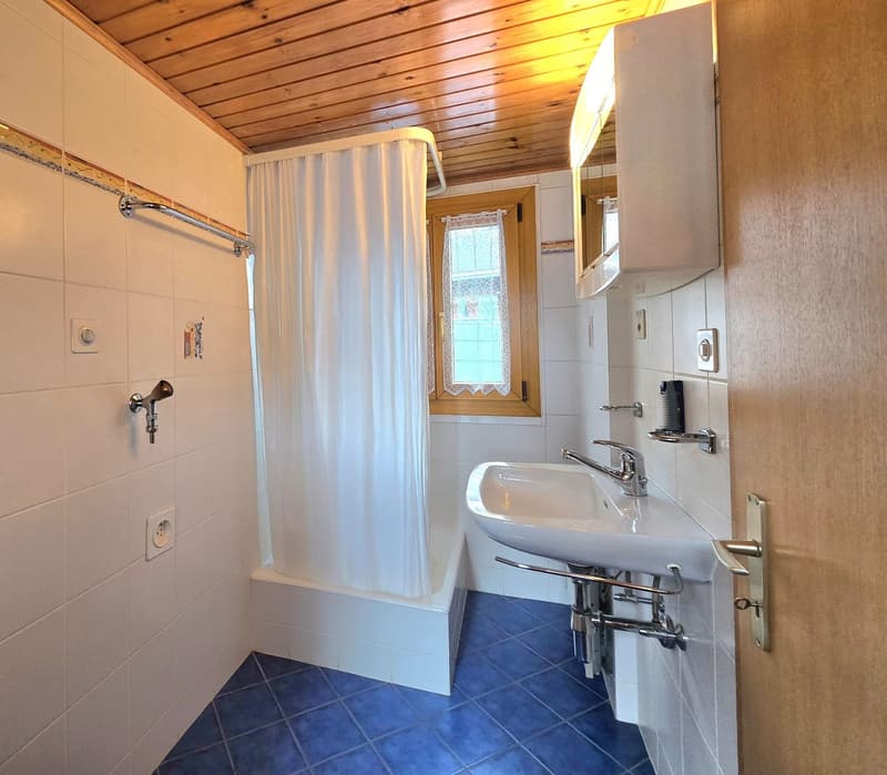 Bathroom with white tiles, blue tiles on the floor, white bathtub, white sink, wooden ceiling, window with white curtains