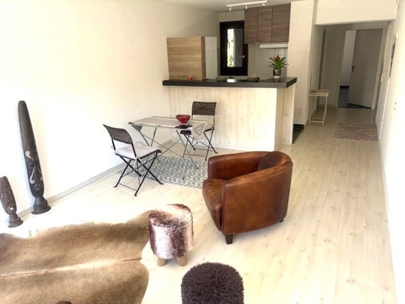 Open plan living and dining area with a kitchen, wooden floor, white walls, a table with chairs, a brown leather armchair, a rug, and a potted plant on the counter.
